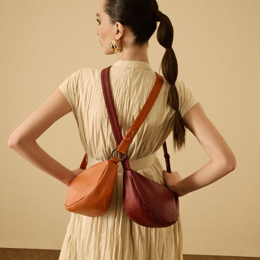 A woman wearing a cream pleated dress stands with her back to the camera, wearing two leather shoulder bags in orange and burgundy with matching straps.