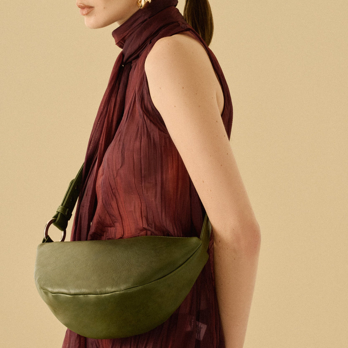 A model in a pleated burgundy dress is shown from the side, wearing a green leather crescent-shaped shoulder bag. The background is a solid, warm beige studio backdrop.