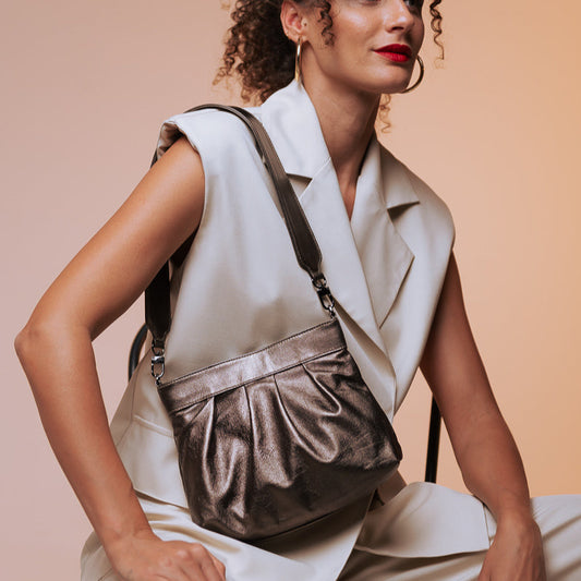 Woman wearing a white sleeveless top and beige pants, holding a brown leather handbag against a beige background.