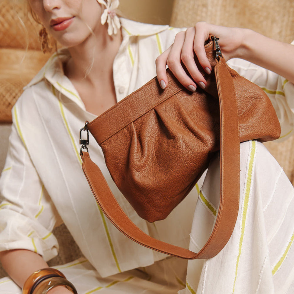 A woman in a cream striped shirt holds a brown pebbled leather shoulder bag with a matching strap, posed against a warm beige background.