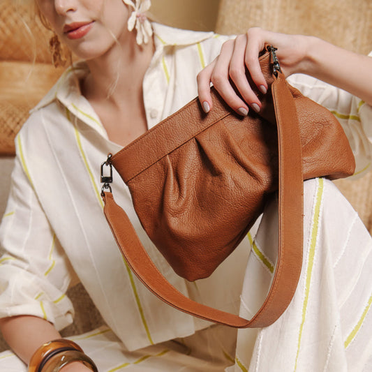 A woman in a cream striped shirt holds a brown pebbled leather shoulder bag with a matching strap, posed against a warm beige background.