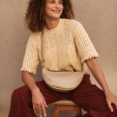 Woman sitting on a stool wearing a beige knitted top and maroon pants against a beige background