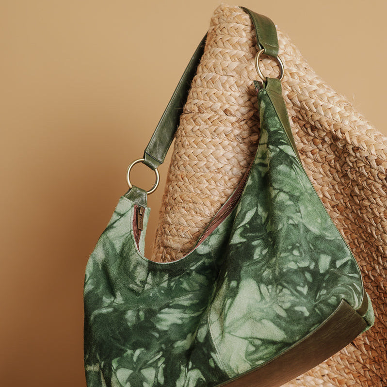 A green and white tie-dye patterned shoulder bag with dark green leather trim, resting on a woven straw chair against a warm tan background.