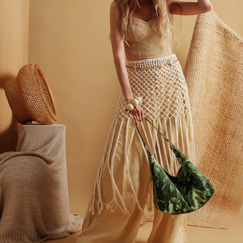 A woman models a bohemian-style outfit featuring a cream crochet halter top, a matching cream macrame skirt with fringe, a large woven straw hat, and a green patterned shoulder bag, set against a warm beige background.