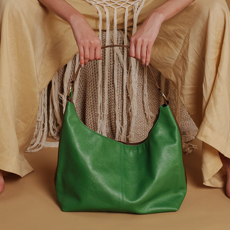 A woman seated on a woven chair wearing beige boho-style clothing and holding a bright green leather hobo bag in front of her.