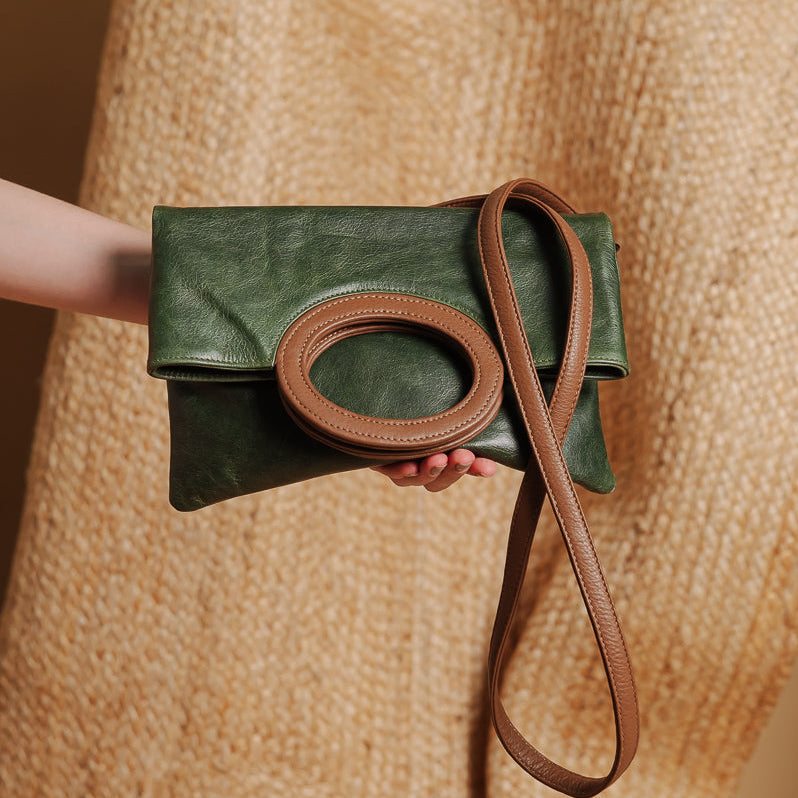 A dark green leather clutch bag with a brown leather oval handle and matching shoulder strap, held by a person wearing a beige knit sweater.
