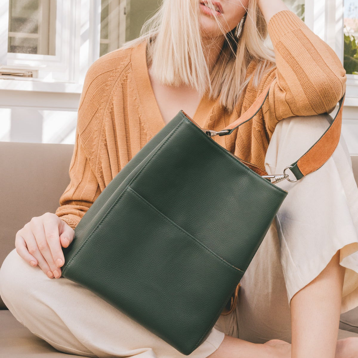 A woman in a rust-colored cardigan and cream pants sits on a beige sofa holding a dark green leather shoulder bag with a tan strap.