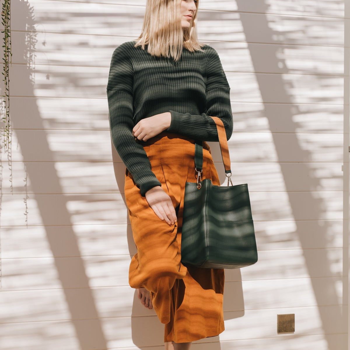 A woman stands barefoot against a white paneled wall with leaf shadows, wearing a dark green ribbed knit sweater and an orange skirt, while carrying a dark green striped tote bag.
