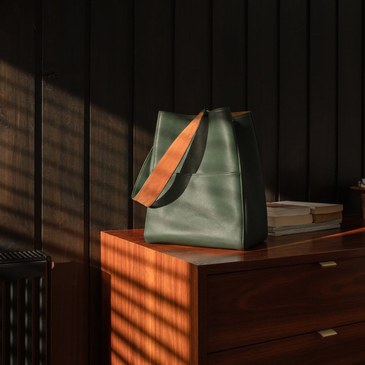A dark green leather tote bag with a contrasting orange-brown shoulder strap, resting on a wooden chest in a room with dramatic, shadowed sunlight.